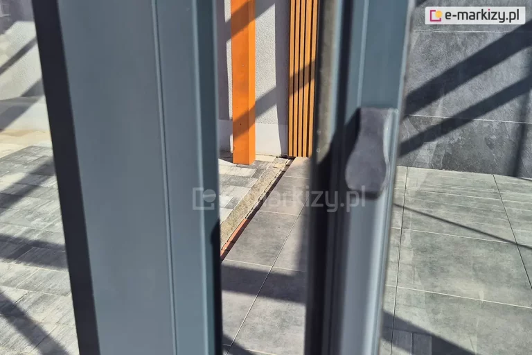 Zdjęcie przedstawia zbliżenie na pionowy profil boczny systemu przesuwnego Sotro, który służy do domykania szklanych skrzydeł. Na pierwszym planie widoczny jest profil aluminiowy w kolorze matowego antracytu z zamontowanym na nim opcjonalnym, pionowym pochwytem o ergonomicznym kształcie. Konstrukcja profilu zamykającego została zaprojektowana tak, aby stabilnie trzymać krawędź przezroczystej tafli szkła. W prześwicie między rozsuniętymi panelami oraz w tle widoczna jest nowoczesna aranżacja tarasu z podłogą wyłożoną dużymi, szarymi płytami oraz fragmentami pionowych lameli dekoracyjnych w kolorze naturalnego drewna. Gra świateł i cieni na posadzce podkreśla transparentność przeszkleń oraz precyzyjne dopasowanie elementów aluminiowych systemu.