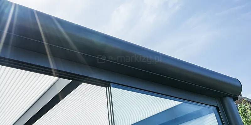 Widok od dołu na zaokrągloną rynnę przednią w zadaszeniu Lunaro Bosco Deponti w kolorze antracytowym, na której widoczne są promienie świecącego z lewej strony słońca. W tle widoczne także niebieskie niebo.