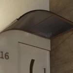 Zamocowany nad drzwiami w narożniku u zbiegu ścian daszek jednostronnie łukowy w kolorze brązowym z wypełnieniem w kolorze dymnym. Płaska storna daszka dotyka ceglanej ściany, Lewy zakrzywiony bok z rynną osłania białe drzwi w jasnej ścianie. Pod daszek widoczne cyfry 1 6 oznaczające numer domu oraz mała kwadratowa zapalona lampka.