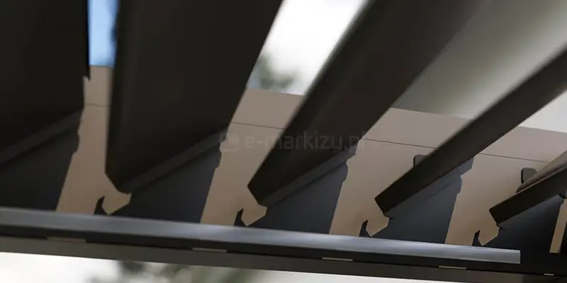 Zbliżenie na detale techniczne ruchomego zadaszenia pergoli Marlo, przedstawiające aluminiowe lamele dachu w pozycji półotwartej. Zdjęcie ukazuje mechanizm mocowania poszczególnych piór do ramy konstrukcyjnej za pomocą systemowych uchwytów. Lamele mają profilowany kształt z wyraźnym przetłoczeniem na krawędzi, co sugeruje obecność systemu odprowadzania wody deszczowej. Kolorystyka elementów jest antracytowa lub taupe, kontrastująca z jasnym tłem nieba widocznym w szczelinach między lamelami. Perspektywa od dołu pozwala zaobserwować precyzję spasowania elementów ruchomych oraz solidność aluminiowych profili tworzących dach bioklimatyczny.