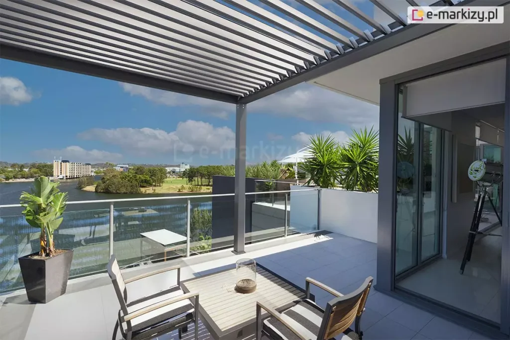 Pergola ogrodowa SB350 Aluprof idealna do samodzielnego montażu zamontowana na tarasie dachowym z pięknym widokiem na rzekę i niebo. Pod pergolą na tarasie ustawiono stolik z dwoma krzesłami oraz roślinę zieloną w dużej grafitowej donicy, na trasie obok widoczne kilka palm.
