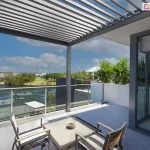 Pergola ogrodowa SB350 Aluprof idealna do samodzielnego montażu zamontowana na tarasie dachowym z pięknym widokiem na rzekę i niebo. Pod pergolą na tarasie ustawiono stolik z dwoma krzesłami oraz roślinę zieloną w dużej grafitowej donicy, na trasie obok widoczne kilka palm.