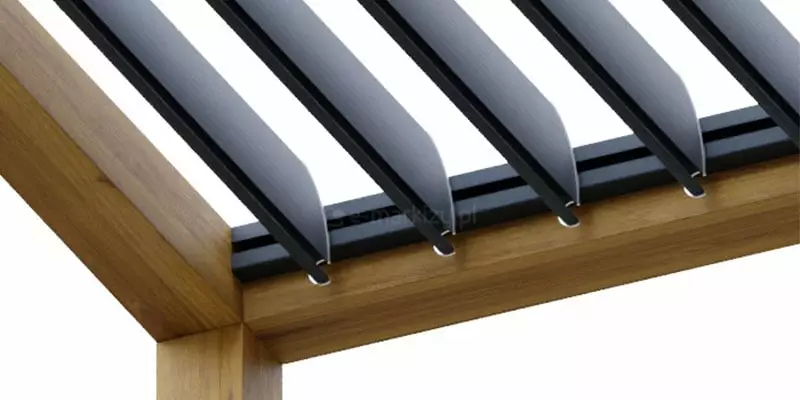 Zbliżenie na detale konstrukcyjne nowoczesnego dachu lamelowego SB400MD do pergoli, które ukazuje sposób montażu ruchomych aluminiowych lameli w ramie wykonanej z litego drewna o naturalnym odcieniu i wyraźnej strukturze usłojenia. Ciemne, malowane proszkowo profile piór dachowych są osadzone w prowadnicach zintegrowanych z drewnianym nadprożem, przy czym widoczne są precyzyjnie wykonane punkty obrotowe oraz elementy mocujące, które umożliwiają płynną regulację kąta nachylenia lameli w celu kontroli nasłonecznienia oraz przepływu powietrza. Drewniana rama nośna, łącząca pionowy słup z poziomą belką konstrukcyjną, stanowi wizualny kontrast dla minimalistycznej, surowej estetyki aluminiowego dachu, podkreślając wysoką jakość wykonania oraz solidność połączeń poszczególnych komponentów systemu zadaszenia. Kąt ujęcia pozwala na dokładną analizę profilu samych piór, które posiadają delikatne przetłoczenia wzmacniające ich sztywność, oraz na obserwację sposobu, w jaki system mechaniczny jest ukryty wewnątrz konstrukcji ramy, co zapewnia czystą i estetyczną linię całego zadaszenia tarasowego nawet w pozycji otwartej.
