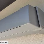 Techniczne zbliżenie od dołu na zamkniętą markizę tarasową Moderno ukrytą w pełnej kasecie osłonowej. Zdjęcie przedstawia prawy skrajny profil konstrukcji, która została zamontowana ściennie we wnęce pod zadaszeniem. Aluminiowa kaseta o nowoczesnym, kubistycznym kształcie z prostymi liniami i zaokrąglonymi narożnikami jest w pełni domknięta, co oznacza, że belka frontowa idealnie przylega do profilu głównego, zabezpieczając tkaninę oraz ramiona przed wpływem warunków atmosferycznych. Cała konstrukcja wykończona jest matowym, strukturalnym lakierem w kolorze jasnoszarym lub srebrnym. Na bocznej, płaskiej pokrywie kasety, w jej prawym dolnym rogu, widnieje mały, prostokątny emblemat z logotypem hiszpańskiego producenta komponentów Llaza. Pod kasetą, w dolnej części profilu, widoczny jest wpuszczany łeb śruby montażowej. Tłem dla urządzenia jest zewnętrzna elewacja budynku wykończona tynkiem strukturalnym typu baranek, przy czym pionowa ściana po prawej stronie jest jasnokremowa i oświetlona promieniami słońca, natomiast górna część wnęki po lewej stronie pozostaje w głębokim, ciemnoszarym cieniu.