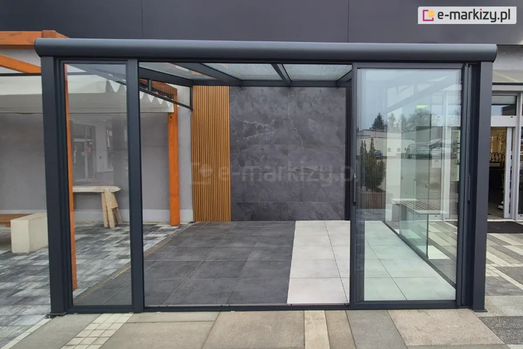 Zdjęcie prezentuje ekspozycję zewnętrznej zabudowy tarasowej w stylu loftowym, wykorzystującą system drzwi szklanych przesuwnych Sotro. Konstrukcja zadaszenia oraz ramy przeszkleń wykonane są z profili aluminiowych o matowym wykończeniu w kolorze ciemnoantracytowym. Frontowa ściana składa się z szerokich, przezroczystych paneli szklanych ujętych w cienkie obramowania, z których centralny segment pozostaje otwarty, ukazując wnętrze tarasu. Wewnątrz strefy wypoczynkowej zastosowano nowoczesne materiały wykończeniowe: tylną ścianę pokrywają wielkoformatowe płyty o teksturze ciemnoszarego kamienia, sąsiadujące z dekoracyjnym panelem z pionowych, drewnianych lameli. Posadzka pod zadaszeniem jest podzielona na dwie strefy kolorystyczne – ciemnoszarą oraz jasnobeżową – wykonane z gładkich płyt tarasowych, które kontrastują z zewnętrzną nawierzchnią z szarej kostki brukowej. Całość tworzy minimalistyczną i funkcjonalną przestrzeń, integrującą nowoczesne wzornictwo z ochroną przed warunkami atmosferycznymi.