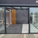 Zdjęcie prezentuje ekspozycję zewnętrznej zabudowy tarasowej w stylu loftowym, wykorzystującą system drzwi szklanych przesuwnych Sotro. Konstrukcja zadaszenia oraz ramy przeszkleń wykonane są z profili aluminiowych o matowym wykończeniu w kolorze ciemnoantracytowym. Frontowa ściana składa się z szerokich, przezroczystych paneli szklanych ujętych w cienkie obramowania, z których centralny segment pozostaje otwarty, ukazując wnętrze tarasu. Wewnątrz strefy wypoczynkowej zastosowano nowoczesne materiały wykończeniowe: tylną ścianę pokrywają wielkoformatowe płyty o teksturze ciemnoszarego kamienia, sąsiadujące z dekoracyjnym panelem z pionowych, drewnianych lameli. Posadzka pod zadaszeniem jest podzielona na dwie strefy kolorystyczne – ciemnoszarą oraz jasnobeżową – wykonane z gładkich płyt tarasowych, które kontrastują z zewnętrzną nawierzchnią z szarej kostki brukowej. Całość tworzy minimalistyczną i funkcjonalną przestrzeń, integrującą nowoczesne wzornictwo z ochroną przed warunkami atmosferycznymi.