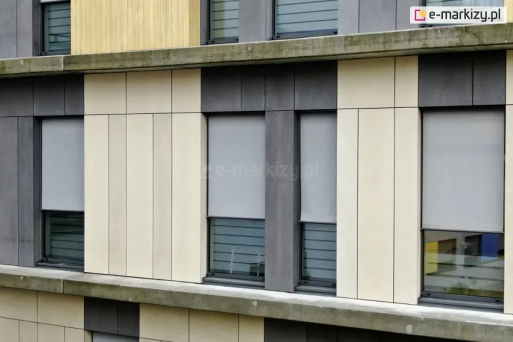 Nowoczesna elewacja budynku biurowego lub mieszkalnego wykonana z pionowych paneli w kolorze piaskowym oraz ciemnoszarym. Na fasadzie widoczne są cztery okna wyposażone w zewnętrzne refleksole 120 Selt. z tkaniną screen w kolorze szarym. Rolety są częściowo opuszczone do około 3/4 wysokości okna, odsłaniając dolne przeszklenia, w pasy matowego i przejrzystego szkła. Tkanina rolet charakteryzuje się drobnym splotem zapewniającym ochronę przeciwsłoneczną przy zachowaniu częściowej przezierności. Systemy osłonowe są zintegrowane z elewacją, z widocznymi prowadnicami bocznymi w kolorze ciemnoszarym, dopasowanymi do stolarki okiennej i paneli fasadowych. Całość kompozycji podkreśla rytm i geometryczny podział nowoczesnej architektury.