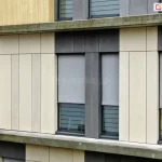 Nowoczesna elewacja budynku biurowego lub mieszkalnego wykonana z pionowych paneli w kolorze piaskowym oraz ciemnoszarym. Na fasadzie widoczne są cztery okna wyposażone w zewnętrzne refleksole 120 Selt. z tkaniną screen w kolorze szarym. Rolety są częściowo opuszczone do około 3/4 wysokości okna, odsłaniając dolne przeszklenia, w pasy matowego i przejrzystego szkła. Tkanina rolet charakteryzuje się drobnym splotem zapewniającym ochronę przeciwsłoneczną przy zachowaniu częściowej przezierności. Systemy osłonowe są zintegrowane z elewacją, z widocznymi prowadnicami bocznymi w kolorze ciemnoszarym, dopasowanymi do stolarki okiennej i paneli fasadowych. Całość kompozycji podkreśla rytm i geometryczny podział nowoczesnej architektury.