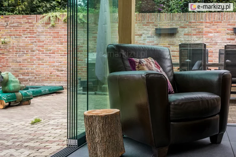 Widok z wnętrza tarasu na system bezramowych szyb przesuwnych Auris w konfiguracji wielotorowej. Na pierwszym planie widoczny jest profil boczny zsuniętych tafli przezroczystego szkła hartowanego, które spoczywają na dolnej, niskoprogowej prowadnicy w kolorze antracytowym. Obok przeszklenia stoi elegancki fotel skórzany w kolorze czarnym z dekoracyjną poduszką oraz rustykalny stolik kawowy wykonany z naturalnego pnia drzewa. Podłoże wewnątrz wykonane jest z dużych, ciemnoszarych płyt tarasowych. Przez szklaną ścianę widoczny jest dziedziniec wyłożony kostką brukową, otoczony murem z czerwonej cegły, przy którym znajduje się niska roślinność ogrodowa.