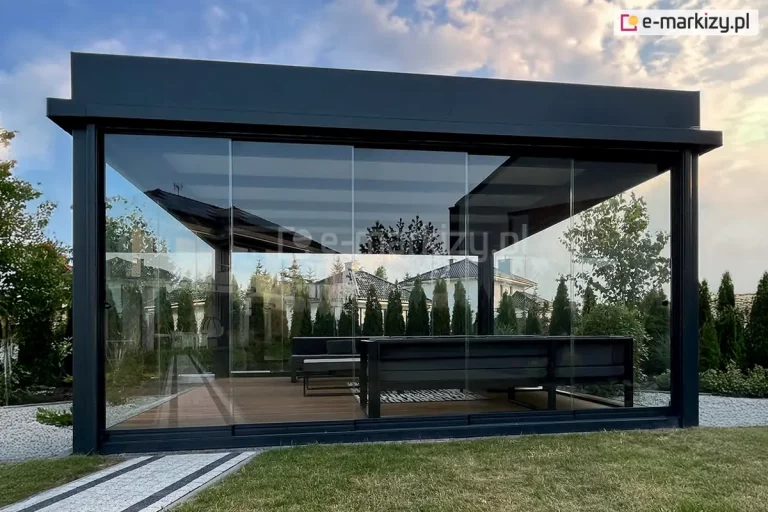 Nowoczesna, wolnostojąca pergola ogrodowa z przeszkleniem całkowitym. Konstrukcja wykonana z ciemnych, aluminiowych profili, wyposażona w przeszklone ściany boczne i frontowe z bezramowymi, przesuwnymi panelami szklanymi model Miros. Wnętrze pergoli zawiera zestaw mebli ogrodowych z kanapą narożną i stołem. Na zewnątrz widać zadbany trawnik, biały żwirowy chodnik i szpaler gęsto rosnących tui. W tle widoczne są dachy i elewacje sąsiednich domów oraz częściowo zachmurzone niebo o zmierzchu.