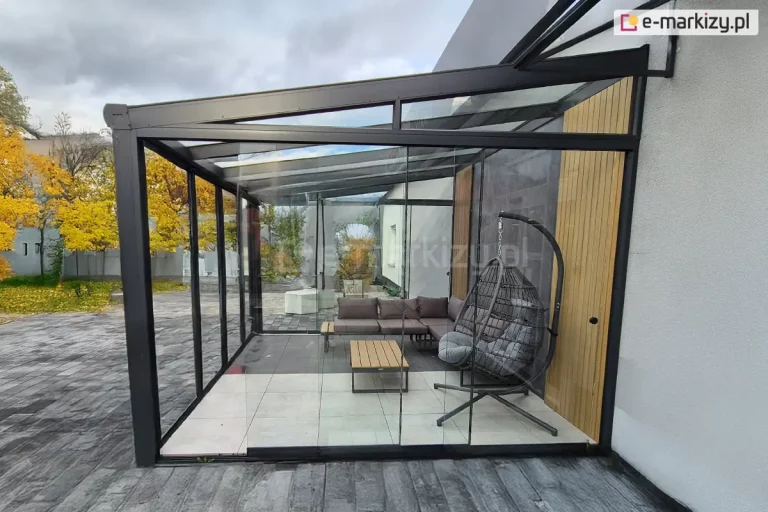 Zdjęcie przedstawia zewnętrzny ogród zimowo-jesienny wykonany z wykorzystaniem szklanych ścian systemu Miros. Konstrukcja tworzy nowoczesną, przeszkloną przestrzeń przy tarasie, chroniącą użytkowników przed wiatrem i chłodem. W pełni przezroczyste panele szklane biegną od podłoża do sufitu, osadzone w aluminiowych profilach i prowadnicach, dzięki czemu wnętrze pozostaje jasne i otwarte wizualnie. W środku widoczne są meble wypoczynkowe – stolik oraz krzesła z poduchami – ustawione na tle roślin i dekoracji sezonowych. Otaczająca zieleń oraz jesienne barwy liści tworzą przytulny, naturalny klimat. Konstrukcja Miros pełni funkcję ochronną i estetyczną, pozwalając korzystać z tarasu przez większą część roku mimo chłodniejszych warunków.