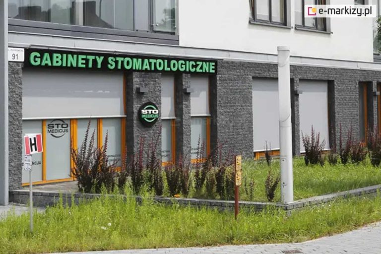 Zdjęcie przedstawia elewację mieszczącego się na parterze piętrowego budynku lokalu usługowego, w którym mieszczą się gabinety stomatologiczne. Budynek posiada nowoczesną fasadę wyłożoną ciemnoszarym kamieniem dekoracyjnym o nieregularnej fakturze. W witrynach okiennych zamontowano zewnętrzne refleksole XXL z tkaniną typu screen w kolorze jasnoszarym. Rolety są częściowo opuszczone, zasłaniając górne partie dużych przeszkleń o pomarańczowych profilach ramowych. Nad głównym wejściem oraz oknami znajduje się długi, czarny szyld z zielonym, podświetlanym napisem „GABINETY STOMATOLOGICZNE”. Na elewacji zamontowano również okrągły kaseton z logo placówki medycznej oraz nowoczesne kinkiety oświetleniowe skierowane w dół. Przed budynkiem znajduje się pas zieleni z niskimi krzewami o ciemnoczerwonych liściach oraz trawnik, oddzielone od chodnika niskim murkiem. Widoczny jest także fragment białego słupa oświetleniowego oraz znak informacyjny na słupku.