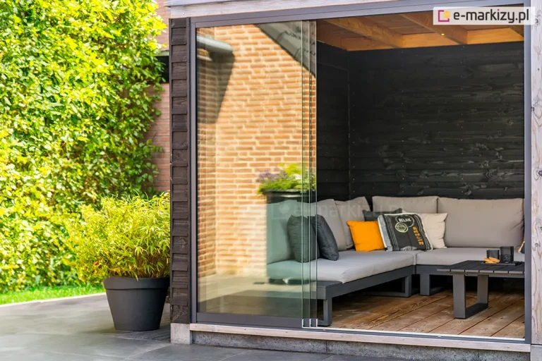 Nowoczesny kącik wypoczynkowy w altanie ogrodowej, zabezpieczony systemem bezramowych szklanych ścian przesuwnych Auris. Zdjęcie przedstawia narożną część konstrukcji z profilami w kolorze antracytowym, gdzie trzy tafle szkła hartowanego są częściowo zsunięte na bok, otwierając wnętrze na taras. W środku znajduje się minimalistyczna, szara sofa modułowa z kolorowymi poduszkami (żółtą i dekoracyjną z napisem) oraz niski, czarny stolik kawowy na drewnianej podłodze. Ściany wewnętrzne altany wyłożone są deskami opalonymi na czarno. Na zewnątrz, obok szklanej zabudowy, stoi duża czarna donica z gęstym bambusem, a w odbiciu szyb widoczna jest nasłoneczniona elewacja z czerwonej cegły. W dalszej perspektywie widoczny zielony żywopłot.