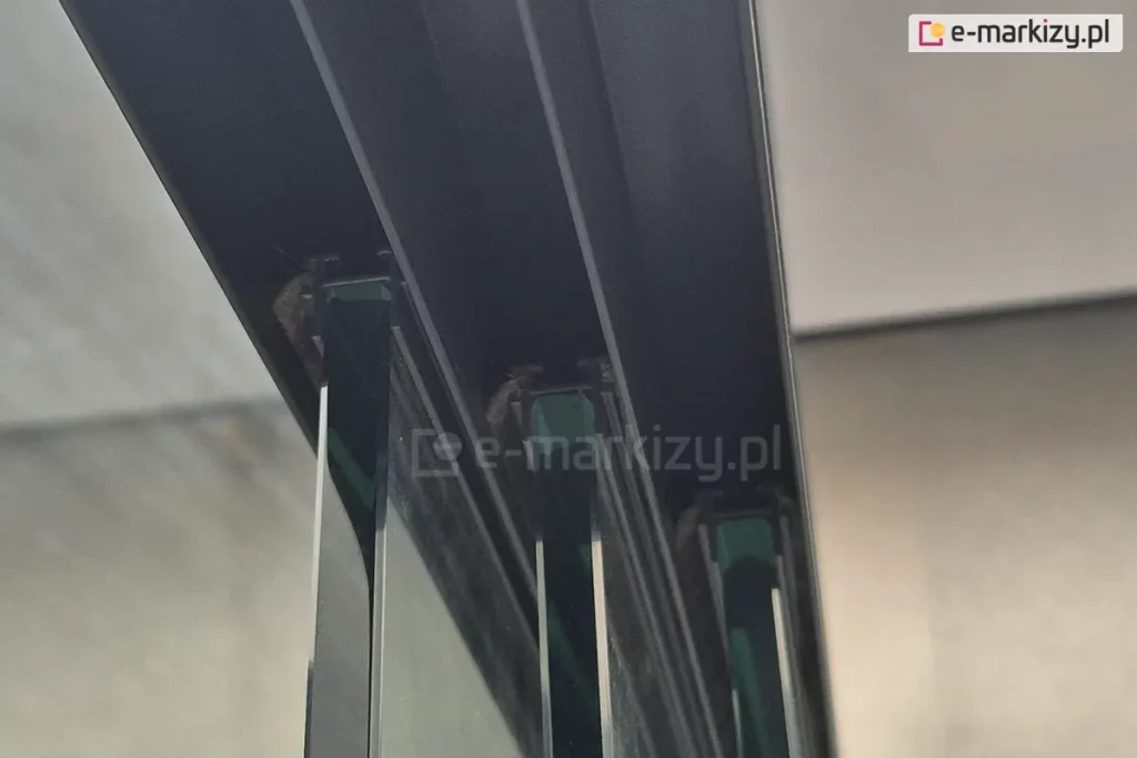 Zbliżenie na górną prowadnicę systemu przesuwnego do szkła, zamontowaną w konstrukcji aluminiowej. Wewnątrz toru znajdują się szczotki uszczelniające, pomiędzy którymi prowadzone są pionowe tafle szkła o lekko zielonym kolorze w przekroju. Widoczne są krawędzie kilku równolegle osadzonych paneli szklanych, prawdopodobnie będących częścią zabudowy tarasowej lub ogrodowej. Na tle rozmyte ściany i sufit z płyt o jasnej barwie.