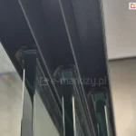 Zbliżenie na górną prowadnicę systemu przesuwnego do szkła, zamontowaną w konstrukcji aluminiowej. Wewnątrz toru znajdują się szczotki uszczelniające, pomiędzy którymi prowadzone są pionowe tafle szkła o lekko zielonym kolorze w przekroju. Widoczne są krawędzie kilku równolegle osadzonych paneli szklanych, prawdopodobnie będących częścią zabudowy tarasowej lub ogrodowej. Na tle rozmyte ściany i sufit z płyt o jasnej barwie.
