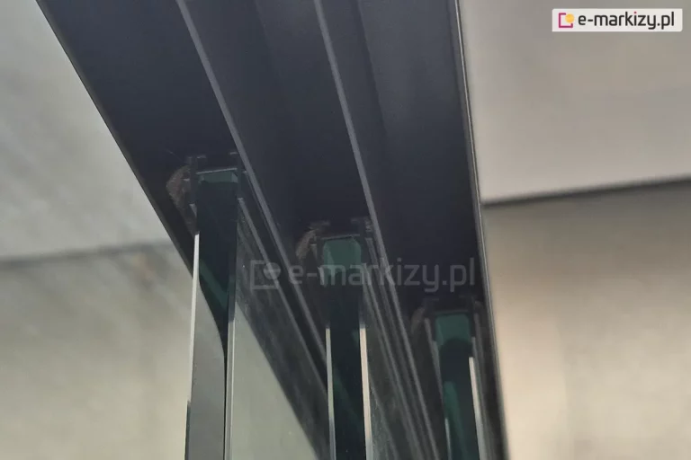 Zbliżenie na górną prowadnicę systemu przesuwnego do szkła, zamontowaną w konstrukcji aluminiowej. Wewnątrz toru znajdują się szczotki uszczelniające, pomiędzy którymi prowadzone są pionowe tafle szkła o lekko zielonym kolorze w przekroju. Widoczne są krawędzie kilku równolegle osadzonych paneli szklanych, prawdopodobnie będących częścią zabudowy tarasowej lub ogrodowej. Na tle rozmyte ściany i sufit z płyt o jasnej barwie.