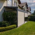 Nowoczesna pergola tarasowa przyścienna Solid o aluminiowej konstrukcji w kolorze ciemnego antracytu, zamontowana do białej elewacji domu jednorodzinnego. Zadaszenie stanowi ruchome, segmentowe poszycie tkaninowe w kolorze kremowym, które na zdjęciu jest rozłożone i zacienia przestrzeń tarasu. Frontowa część pergoli została wyposażona w dwie pionowe rolety zewnętrzne typu screen w kolorze beżowym, przy czym lewa roleta jest całkowicie opuszczona, a prawa podniesiona do połowy wysokości. Konstrukcja opiera się na masywnych słupach o kwadratowym przekroju, a pod nią znajduje się taras wyłożony szarymi płytami gresowymi. Bezpośrednio przy pergoli widoczna jest drewniana kratka ogrodowa gęsto porośnięta winoroślą z owocami oraz niski, formowany żywopłot. W tle rozpościera się zadbany trawnik, wysoki płot z siatki porośnięty bluszczem oraz trzy krzewy ozdobne o stożkowym kształcie. Zdjęcie wykonane w słoneczny dzień, prezentuje pełne możliwości osłony tarasu za pomocą systemów rolet bocznych.