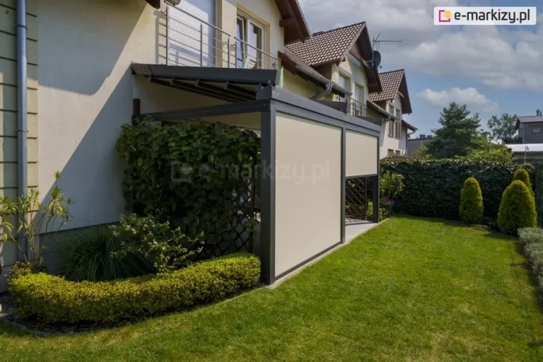 Nowoczesna pergola tarasowa przyścienna Solid o aluminiowej konstrukcji w kolorze ciemnego antracytu, zamontowana do białej elewacji domu jednorodzinnego. Zadaszenie stanowi ruchome, segmentowe poszycie tkaninowe w kolorze kremowym, które na zdjęciu jest rozłożone i zacienia przestrzeń tarasu. Frontowa część pergoli została wyposażona w dwie pionowe rolety zewnętrzne typu screen w kolorze beżowym, przy czym lewa roleta jest całkowicie opuszczona, a prawa podniesiona do połowy wysokości. Konstrukcja opiera się na masywnych słupach o kwadratowym przekroju, a pod nią znajduje się taras wyłożony szarymi płytami gresowymi. Bezpośrednio przy pergoli widoczna jest drewniana kratka ogrodowa gęsto porośnięta winoroślą z owocami oraz niski, formowany żywopłot. W tle rozpościera się zadbany trawnik, wysoki płot z siatki porośnięty bluszczem oraz trzy krzewy ozdobne o stożkowym kształcie. Zdjęcie wykonane w słoneczny dzień, prezentuje pełne możliwości osłony tarasu za pomocą systemów rolet bocznych.