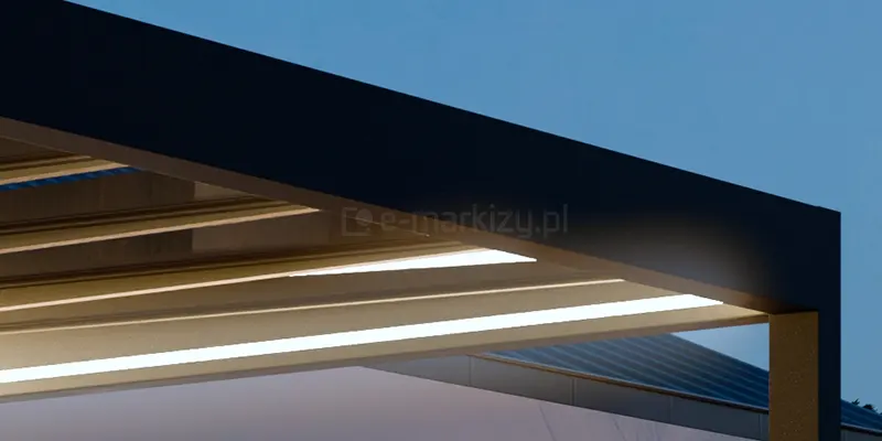 Zbliżenie na detal konstrukcyjny nowoczesnego zadaszenia szklanego Spekta Glass w warunkach wieczornych. Na pierwszym planie widoczny jest narożnik ramy obwodowej wykonanej z grubego profilu aluminiowego w kolorze antracytowym o matowym wykończeniu. Pod konstrukcją dachu zintegrowano system oświetlenia LED w formie szerokich, liniowych pasów świetlnych zamontowanych wzdłuż poprzecznych krokwi. Oświetlenie emituje intensywne, białe światło o chłodnej barwie, które jest skierowane ku dołowi, oświetlając przestrzeń pod zadaszeniem. W tle, pod kątem, widoczne są kolejne sekcje przeszklonego dachu oraz fragment elewacji budynku i pochyłego dachu krytego blachą. Całość ukazana jest na tle ciemniejącego, granatowego nieba, co podkreśla funkcjonalność i estetykę zintegrowanego systemu świetlnego w nowoczesnej architekturze tarasowej.