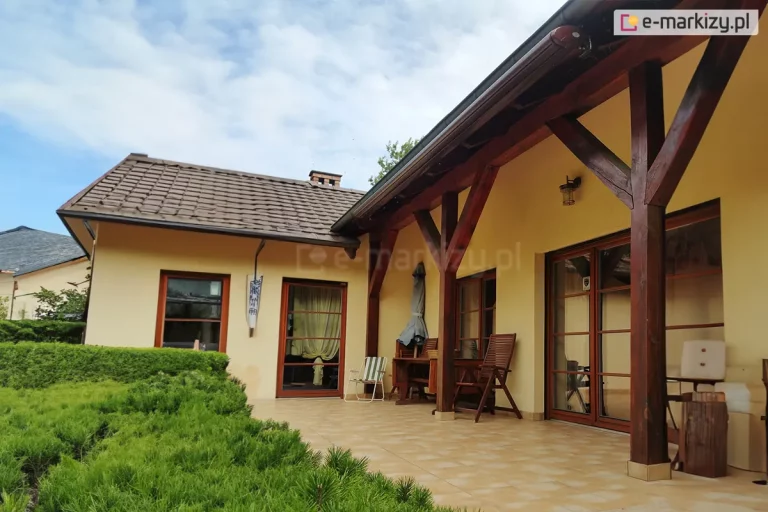 Zdjęcie prezentuje frontową elewację parterowego budynku jednorodzinnego z rozległym tarasem, na którym zamontowana jest zwinięta markiza w pełnej kasecie. Kaseta markizy w kolorze ciemnego brązu znajduje się pod okapem dachu, przylegając do ciemnych, drewnianych belek konstrukcyjnych wspierających zadaszenie tarasu. Ściany budynku wykończone są gładkim tynkiem w kolorze piaskowym lub jasnożółtym. Taras wyłożony jest kwadratowymi płytkami ceramicznymi w odcieniach beżu i jasnego brązu, tworząc spójną całość z kolorystyką elewacji. Na tarasie znajdują się liczne przeszklenia: szerokie drzwi tarasowe z brązowymi ramami oraz mniejsze okno, w których odbija się otoczenie. Przestrzeń wypoczynkowa jest wyposażona w drewniane meble ogrodowe, w tym krzesła i stół, a obok drzwi stoi białe, składane krzesło. Po lewej stronie kadru widoczny jest niski, gęsty żywopłot z iglaków, oddzielający taras od reszty ogrodu. Dach domu pokryty jest brązową dachówką, a z jego połaci wystaje klasyczny komin murowany z cegły. Niebo nad budynkiem jest jasne, z rozproszonymi, białymi chmurami, co nadaje scenie spokojny, popołudniowy charakter. Perspektywa zdjęcia obejmuje całą bryłę tarasu, ukazując dyskretny montaż markizy w pełnej kasecie, która po zwinięciu idealnie komponuje się z elementami stolarki i konstrukcji dachowej.