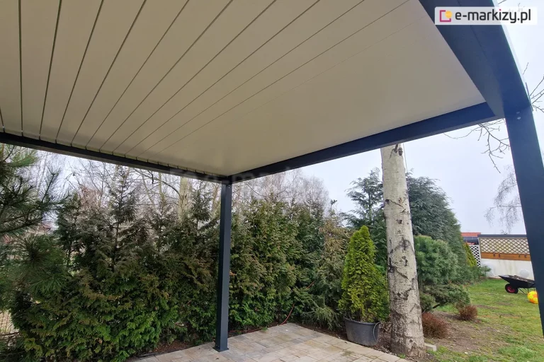 Widok od dołu na szczelnie zamknięte lamele dachowe pergoli SB350 Selt. Białe, aluminiowe panele tworzą jednolitą, gładką płaszczyznę zadaszenia, wspartą na antracytowej ramie i pionowych słupach nośnych. Konstrukcja rzuca cień na taras wyłożony jasną kostką brukową. W tle widoczna jest gęsta roślinność ogrodowa z wysokimi tujami, sosnami oraz pniem brzozy w donicy. W oddali po prawej stronie widać fragment trawnika z taczką ogrodową oraz ozdobną kratkę pergoli drewnianej pod jasnym, zachmurzonym niebem.
