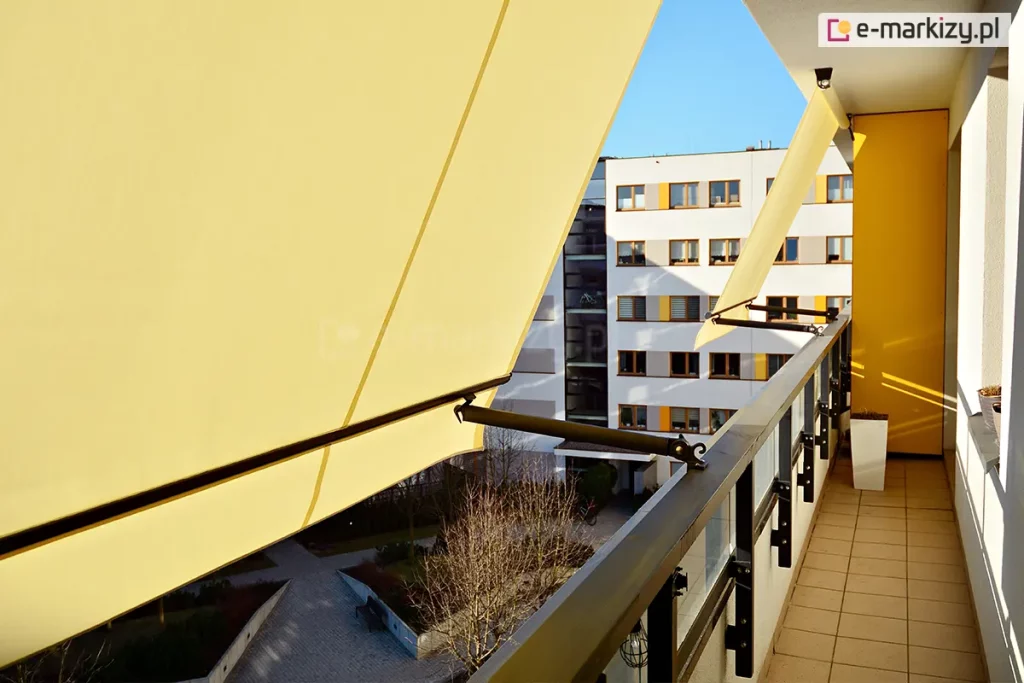 Dwie markizy balkonowe-poręczowe z jasnożółtym poszyciemi antracyowymi ramionami przymocowanymi do barierki wąskiego balkonu wyłożonego płytkami. W tle żółta ściana ogradzająca sąsiedni balkon, a przed nią duża biała kwadratowa donica, w której nie widać roślin. W tle widoczny sąsiedni budynek i alejki pomiędzy blokami.