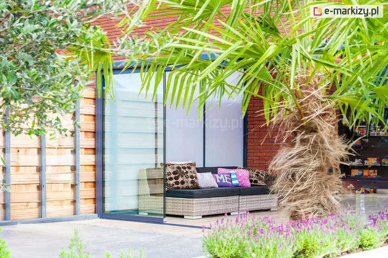 Widok na częściowo zsuniętą bezramową ścianę szklaną systemu Auris, zamontowaną w nowoczesnej altanie ogrodowej. Na pierwszym planie widoczna jest tafla przezroczystego szkła hartowanego o szerokości panelu 104 cm, przesunięta na prowadnicy bocznej. Wewnątrz osłoniętej przestrzeni znajduje się wygodna sofa z szarego technorattanu z ciemnymi siedziskami i ozdobnymi poduszkami. Tło stanowi elewacja z czerwonej cegły oraz białe panele wypełniające tył konstrukcji. Całość aranżacji dopełnia egzotyczna roślinność – liście palmy wchodzące w kadr oraz fioletowe kwiaty lawendy na dole. Konstrukcja ramy utrzymana jest w kolorze antracytowym.