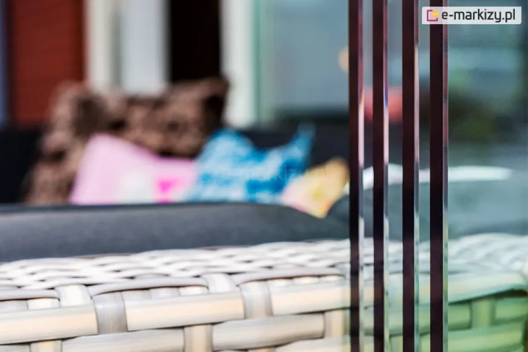 Zbliżenie na pionowe krawędzie zsuniętych paneli szklanych w bezramowym systemie przesuwnym Auris. Zdjęcie wykonane z boku, z płytką głębią ostrości, eksponuje cztery tafle przezroczystego szkła hartowanego o precyzyjnie oszlifowanych i wypolerowanych krawędziach. Panele są ułożone równolegle, prezentując kaskadowy mechanizm składania ściany szklanej. W rozmytym tle widoczne jest wnętrze strefy wypoczynkowej z meblami z technorattanu oraz kolorowymi poduszkami dekoracyjnymi. Detal podkreśla minimalistyczny design i wysoką jakość obróbki szkła, która zapewnia bezpieczeństwo użytkowania oraz estetyczny wygląd zabudowy tarasu.