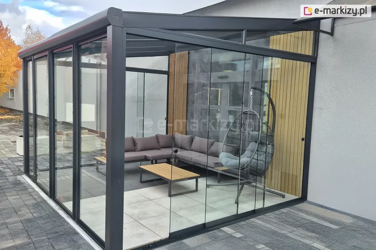 Szklane zadaszenie tarasu z widocznymi na bocznej ścianie szklanymi ścianami przesuwnymi bezramowymi Miros o lekko przesuniętym skrajnym skrzydle. Na skrajnych panelach zamocowane czarne okrągłe klamki. Po lewej stronie, od frontu zadaszenia widoczny ramowy system szyb przesuwnych. Pod zadaszeniem ustawiono niski stolik z drewnianym blatem oraz narożnik wyłożony szaro-brązowymi poduszkami. Obok narożnika duży fotel wiszący na stelażu. W tle, przy jasnej ścianie budynku widoczne drzewo obsypane jesiennymi, złotymi liśćmi.