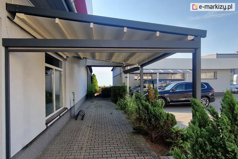 Zdjęcie przedstawia nowoczesną konstrukcję pergoli tarasowej Square zamontowanej przy elewacji budynku, która pełni funkcję zadaszenia nad przejściem wyłożonym kostką brukową. Konstrukcja wspierająca wykonana jest z profili aluminiowych o matowym wykończeniu w kolorze antracytu. Składa się ona z solidnych pionowych słupów nośnych oraz ramy poziomej zamocowanej z jednej strony bezpośrednio do białej ściany budynku, a z drugiej wspartej na zewnętrznych filarach. Zadaszenie stanowi ruchome poszycie tkaninowe w kolorze kremowym lub złamanej bieli, które jest rozpięte na poprzecznych belkach przeciwwietrznych. Materiał układa się w charakterystyczne segmenty i jest zamontowany pod lekkim skosem, co umożliwia swobodny odpływ wody opadowej. Pod zadaszeniem znajduje się chodnik z szarej kostki brukowej, prowadzący wzdłuż ściany z dużym okien o białych ramach. Na widocznej na zdjęciu bocznej ścianie zamocowano poprzeczną belkę łączącą podporę ze ścianą, do której przymocowany został refleksol Zip L Zip U, stanowiący osłonę boczną pergoli. Po prawej stronie konstrukcji widoczny jest pas zieleni z niskimi krzewami iglastymi oraz parking, na którym zaparkowany jest czarny samochód osobowy typu SUV oraz inne pojazdy w tle. W oddali widać fragmenty sąsiednich budynków oraz inne wiaty garażowe. Niebo jest bezchmurne i jasne, co sugeruje słoneczny dzień, a cała scena ukazuje praktyczne zastosowanie pergoli jako osłony przejścia komunikacyjnego przy obiekcie komercyjnym lub mieszkalnym.