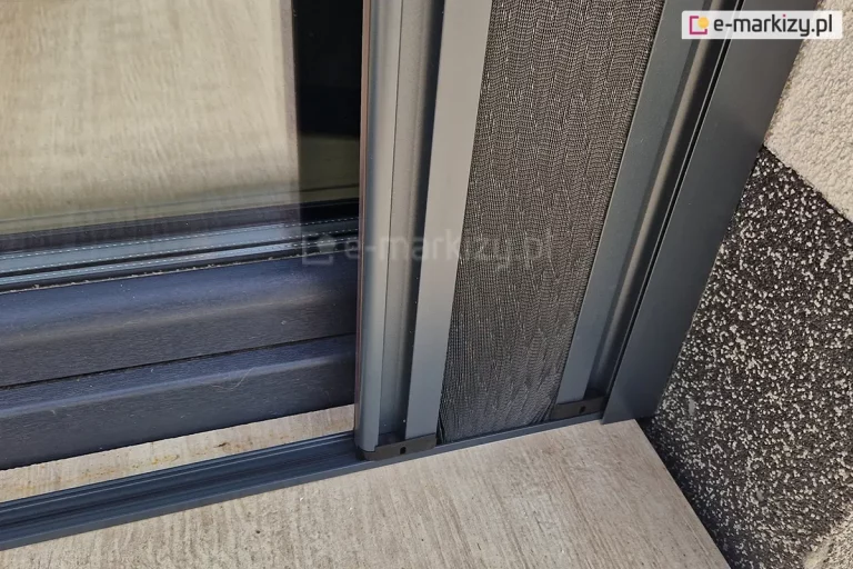 Zwinięty pakiet siatki z profilami bocznymi moskitiery Top Zag zwinięty do ściany o białym i szarym tynku. Próg naklejony na płytki gresowe o drewnianym wzorze, w lekkim oddaleniu od ramy okna o grafitowym kolorze.