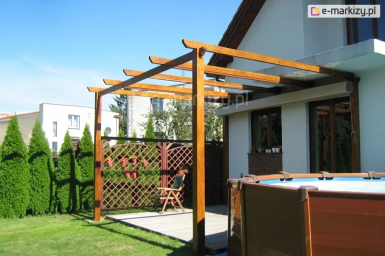 Zdjęcie przedstawia nowoczesną konstrukcję pergoli drewnianej zamontowanej przy elewacji budynku mieszkalnego, pełniącą funkcję zadaszenia tarasu. Konstrukcja wsparta jest na solidnych, pionowych słupach z drewna klejonego w kolorze średniego brązu o wyraźnym usłojeniu. Górną część stanowią równolegle rozmieszczone krokwie z ozdobnie ściętymi końcami, wysuniętymi poza obrys przedniej belki nośnej. Na szczycie konstrukcji widoczny jest zwinięty dach tkaninowy systemu Supro, który w tej pozycji spoczywa w dyskretnej kasecie lub pod osłoną przy samej ścianie budynku, odsłaniając całkowicie przestrzeń nad tarasem. Pod pergolą znajduje się podest wykonany z szarych desek tarasowych. W tle widoczne jest ażurowe ogrodzenie lamelowe z zawieszonymi doniczkami, drewniane krzesło ogrodowe oraz fragment basenu stelażowego o brązowym wykończeniu ścianek. Całość zlokalizowana jest w zadbanym ogrodzie z zielonym trawnikiem i rzędem wysokich tui, w pełnym świetle słonecznym.