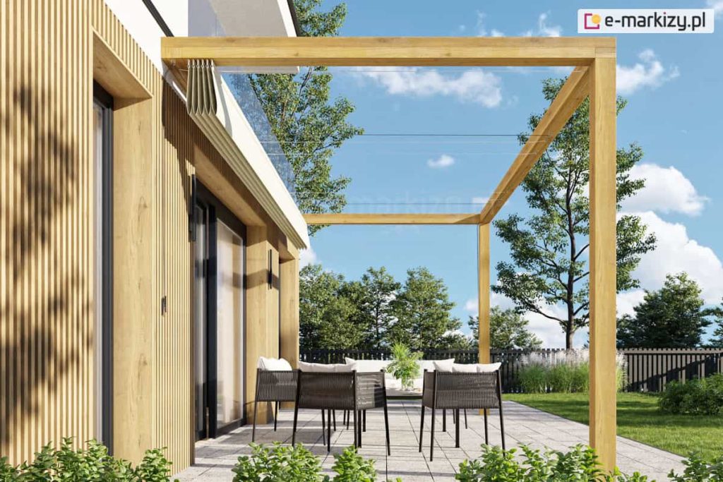 Zdjęcie prezentuje dach rzymski Risto w stanie całkowicie złożonym, zamontowany na drewnianej pergoli przyściennej. Beżowa tkanina zadaszenia jest zwinięta w wąski, estetyczny pakiet podwieszony pod belką poprzeczną przy samej elewacji budynku, co odsłania konstrukcję nośną pergoli oraz błękitne niebo. Przez wolną przestrzeń pod belkami przebiegają cienkie, poziome linki stalowe, po których przesuwa się materiał podczas rozkładania. Konstrukcja zadaszenia wykonana jest z belek o jasnym, naturalnym odcieniu drewna i przylega do białej ściany domu wykończonej pionowymi lamelami. Na tarasie z jasnych płyt betonowych ustawiony jest nowoczesny zestaw wypoczynkowy z ciemnoszarymi fotelami, a w tle widoczny jest zadbany ogród z trawnikiem i drzewami. Perspektywa zdjęcia ukazuje, jak niewielką przestrzeń zajmuje złożony system tkaninowy, nie ograniczając dostępu światła na taras w pogodne dni.