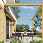 Zdjęcie prezentuje dach rzymski Risto w stanie całkowicie złożonym, zamontowany na drewnianej pergoli przyściennej. Beżowa tkanina zadaszenia jest zwinięta w wąski, estetyczny pakiet podwieszony pod belką poprzeczną przy samej elewacji budynku, co odsłania konstrukcję nośną pergoli oraz błękitne niebo. Przez wolną przestrzeń pod belkami przebiegają cienkie, poziome linki stalowe, po których przesuwa się materiał podczas rozkładania. Konstrukcja zadaszenia wykonana jest z belek o jasnym, naturalnym odcieniu drewna i przylega do białej ściany domu wykończonej pionowymi lamelami. Na tarasie z jasnych płyt betonowych ustawiony jest nowoczesny zestaw wypoczynkowy z ciemnoszarymi fotelami, a w tle widoczny jest zadbany ogród z trawnikiem i drzewami. Perspektywa zdjęcia ukazuje, jak niewielką przestrzeń zajmuje złożony system tkaninowy, nie ograniczając dostępu światła na taras w pogodne dni.