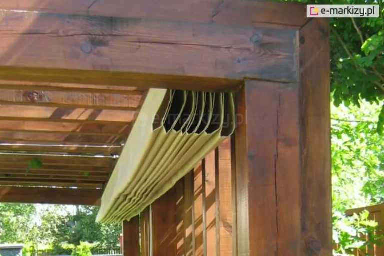 Zbliżenie na pakiet zwiniętej tkaniny dachowej systemu Supro, zamontowany na konstrukcji pergoli wykonanej z grubych belek drewna klejonego w ciemnobrązowym odcieniu. Widoczny jest pakiet materiału w jasnobeżowym kolorze, który został złożony w równomierne fałdy i ściągnięty do jednej z krawędzi zadaszenia. Tkanina dachu rzymskiego jest podwieszona pod poprzecznymi krokwiami za pomocą systemu prowadnic, co umożliwia jej płynne rozsuwanie i zsuwanie. Drewniana konstrukcja wykazuje naturalne cechy materiału, takie jak sęki i pęknięcia strukturalne, co podkreśla solidny charakter zabudowy. W tle, pod zadaszeniem, widać fragment ażurowych przesłon bocznych oraz naturalną zieleń ogrodową, co obrazuje sposób montażu systemu wewnątrz ramy pergoli.