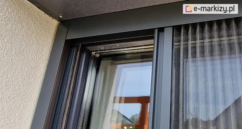 Listwa górna i prowadnica boczna stanowiące narożnik moskitiery plisowanej z szarą siatką i antracytową konstrukcją, zamocowane do prowadnic rolety zewnętrznej we wnęce okna o białym tynku strukturalnym.