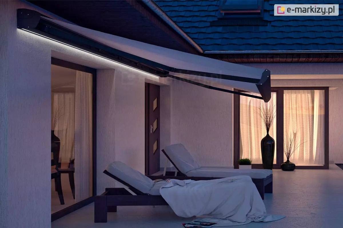 Zdjęcie przedstawia nowoczesną markizę Corsica zamontowaną na elewacji piętrowego domu jednorodzinnego w porze wieczornej. Markiza Corsica jest w pełni rozwinięta, a jej konstrukcja opiera się na bocznych ramionach przegubowych pomalowanych na ciemny, prawdopodobnie antracytowy kolor, który kontrastuje z jasnym poszyciem materiałowym. Tkanina markizy ma kolor biały lub bardzo jasnoszary i jest napięta nad tarasem, tworząc zadaszenie. Charakterystycznym elementem widocznym na zdjęciu jest zintegrowane oświetlenie LED w postaci długiej listwy świetlnej zamontowanej wzdłuż kasety markizy przy samej ścianie budynku. Światło o chłodnej barwie rzuca wyraźny blask na górną część elewacji oraz pod spód zadaszenia, doświetlając przestrzeń wypoczynkową. Pod markizą znajdują się dwa nowoczesne leżaki ogrodowe z ciemnymi ramami i jasnymi materacami, na których niedbale ułożono białe koce lub ręczniki. W tle widoczna jest jasna, strukturalna elewacja budynku z dużymi przeszkleniami oraz drzwiami wejściowymi o nowoczesnym wzornictwie. Przez okna tarasowe przebija ciepłe, żółte światło z wnętrza domu, gdzie dostrzec można elementy wystroju, takie jak wysoki, ciemny wazon z ozdobnymi trawami. Nawierzchnia tarasu jest gładka i jasna, odbijająca sztuczne światło płynące z markizy oraz z wnętrza pomieszczeń. Dach budynku pokryty jest ciemną dachówką, a cała scena utrzymana jest w chłodnej, wieczornej kolorystyce, która podkreśla funkcjonalność oświetlenia markizy Corsica.