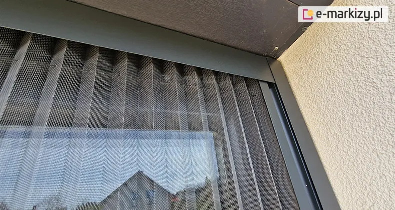 Prawy narożnik moskitiery plisowanej Top Zag z charakterystycznym łączeniem profilu bocznego z górnym, wykonanych w kolorze antracytu z szarą siatką na oknie, w którym odbija się sąsiadujący dom.