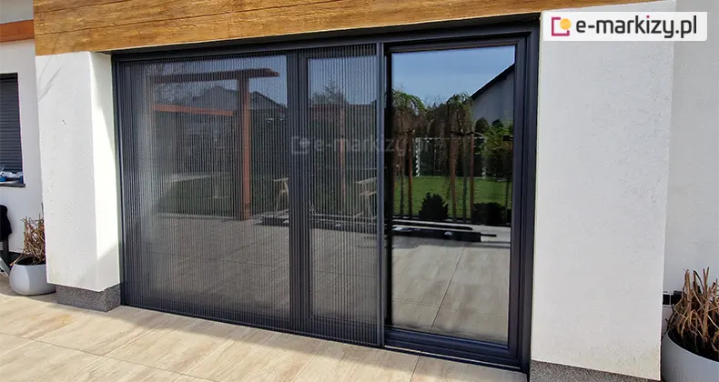 Duże okno tarasowe o antracytowej ramie osłonięte częściowo moskitierą plisowaną Top Zag, zamontowaną do ramy okna. Okno otoczone białą elewacją oraz drewnianym wykończeniem górnej części, Na tarasie duże jasne płytki oraz dwie donice z zasuszonymi roślinami.