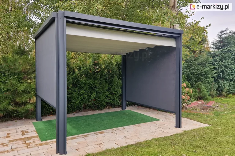 Wolnostojąca pergola tarasowa Tako o antracytowej konstrukcji aluminiowej, wyposażona w jasny, segmentowy dach tkaninowy oraz dwie rozwinięte boczne rolety typu refleksol w kolorze ciemnoszarym. Pergola jest ustawiona w ogrodzie na podłożu z jasnej kostki brukowej, na której położono pas sztucznej trawy w kolorze intensywnej zieleni. Zadaszenie jest w całości rozłożone, tworząc falowaną powierzchnię. Konstrukcja wsparta na czterech narożnych słupach znajduje się na tle gęstych, zielonych krzewów iglastych oraz drzew liściastych z żółknącymi liśćmi. Po prawej stronie widoczne są fragmenty drewnianych belek leżących na trawniku oraz kwiaty ogrodowe.