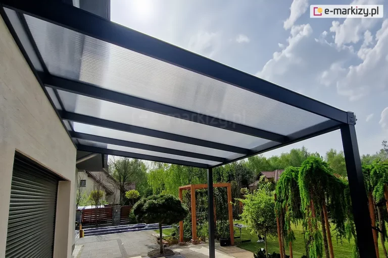 Zdjęcie prezentuje ujęcie z perspektywy żabiej na nowoczesną, przyścienną konstrukcję zadaszenia tarasowego Lunaro w kolorze antracytowym. Aluminiowy stelaż jest zamontowany bezpośrednio do jasnej elewacji budynku i wspiera się na dwóch podporach o kwadratowym przekroju. Dach zadaszenia posiada wypełnienie z półprzezroczystych płyt poliwęglanu komorowego, które wpuszczają miękkie, rozproszone światło. Pod zadaszeniem, na jasnym tarasie, widoczny jest fragment ciemnej rolety zewnętrznej oraz małe drzewko iglaste w donicy. W tle rozciąga się widok na słoneczny ogród z różnorodną roślinnością, obejmujący ozdobne pergole, krzewy oraz drzewa liściaste pod jasnym niebem. Po lewej stronie widoczny jest fragment sąsiedniego domu z charakterystycznym spadzistym dachem.