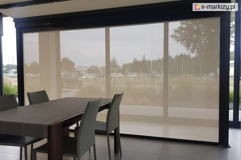 Pomieszczenie biurowe z dużym stołem i czterema krzesłami, utrzymane w szarej kolorystyce, przesłonięte zostało refleksolem ZIP L, który ogranicza ilość słońca wpadającego do pomieszczenia oraz widoczność z zewnątrz. Przez roletę widoczny jest trawnik przed budynkiem oraz znajdujący się nieco dalej parking.