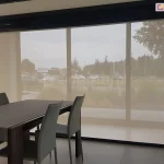 Pomieszczenie biurowe z dużym stołem i czterema krzesłami, utrzymane w szarej kolorystyce, przesłonięte zostało refleksolem ZIP L, który ogranicza ilość słońca wpadającego do pomieszczenia oraz widoczność z zewnątrz. Przez roletę widoczny jest trawnik przed budynkiem oraz znajdujący się nieco dalej parking.