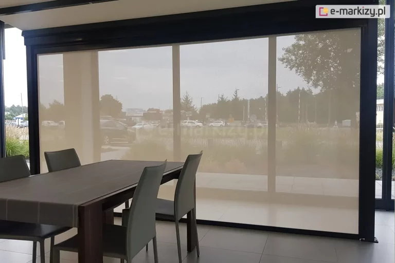 Pomieszczenie biurowe z dużym stołem i czterema krzesłami, utrzymane w szarej kolorystyce, przesłonięte zostało refleksolem ZIP L, który ogranicza ilość słońca wpadającego do pomieszczenia oraz widoczność z zewnątrz. Przez roletę widoczny jest trawnik przed budynkiem oraz znajdujący się nieco dalej parking.