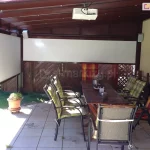 Pergola tarasowa w kolorze brązowym z dwoma refleksolami na boku i na froncie, wyposażonymi w białą tkaninę, Na tarasie z płytek gresowych pod pergolą ustawiono dwa drewniane stoły i wokół nich kilka krzeseł z wygodnymi poduszkami w kratę, Za refleksolem widoczny płot z paneli drewnianych oraz trawnik.