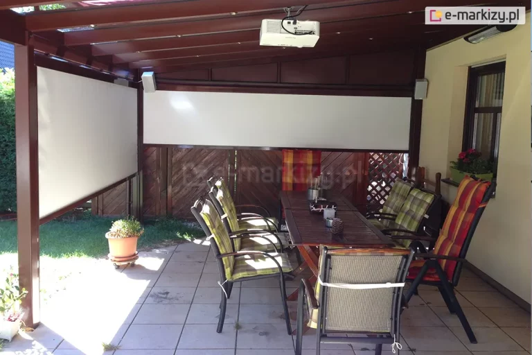 Pergola tarasowa w kolorze brązowym z dwoma refleksolami na boku i na froncie, wyposażonymi w białą tkaninę, Na tarasie z płytek gresowych pod pergolą ustawiono dwa drewniane stoły i wokół nich kilka krzeseł z wygodnymi poduszkami w kratę, Za refleksolem widoczny płot z paneli drewnianych oraz trawnik.