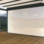 Duże zadaszenie z dachem tkaninowym i ścianką frontową refleksol ZIP z tkaniną przezierną ustawione zostało na drewnianym pustym tarasie. Za metalowymi barierkami w tle widoczne zabudowania, lasy oraz niewysokie góry i niebieskie niebo z kilkoma chmurami.