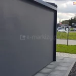 Ściana zadaszenia wykonana z rolety ZIP L skutecznie osłania, co się dzieje na tarasie przed wzrokiem przechodniów. W tle widoczny trawnik, droga z kostki brukowej oraz parking.