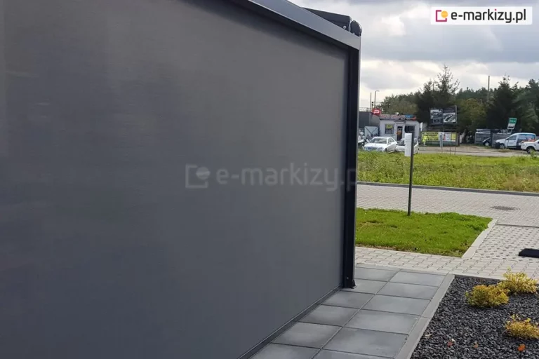 Ściana zadaszenia wykonana z rolety ZIP L skutecznie osłania, co się dzieje na tarasie przed wzrokiem przechodniów. W tle widoczny trawnik, droga z kostki brukowej oraz parking.
