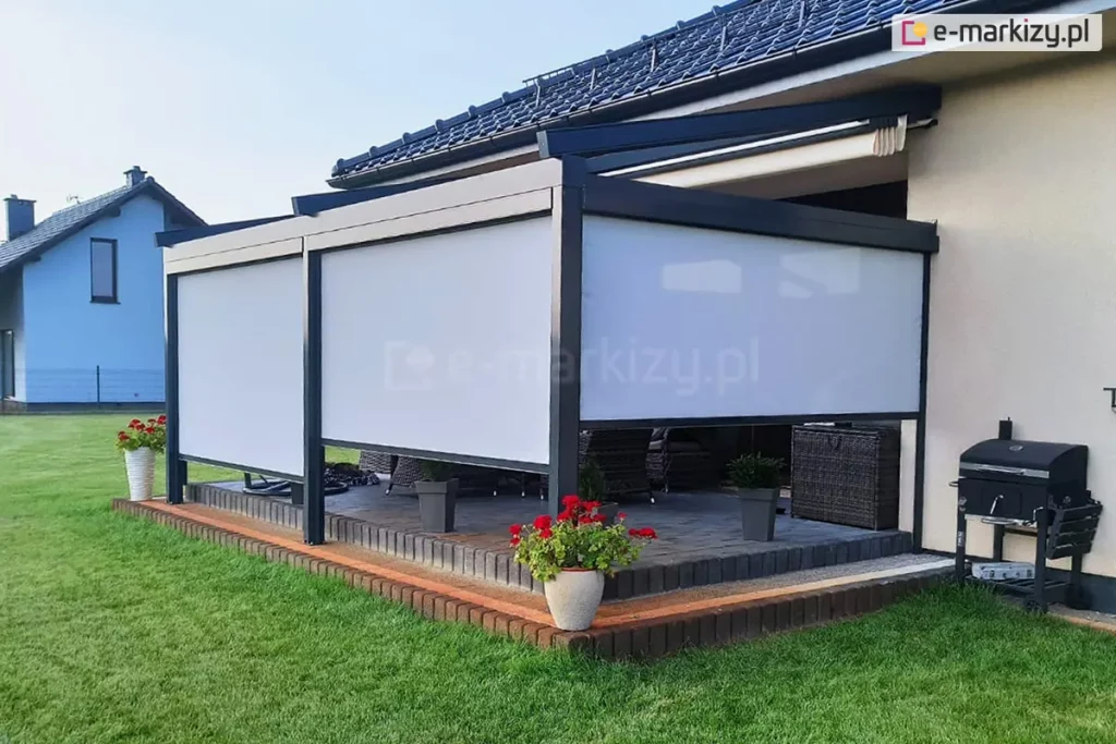 Pergola tarasowa ze zwiniętym dachem tkaninowym i z bocznymi roletami typu refleksole ZIP L wykonanymi z półprzezroczystej tkaniny, zamontowana przy nowoczesnym domu jednorodzinnym. Pod zadaszeniem znajduje się zestaw mebli ogrodowych, a wokół ustawiono donice z kwiatami, po prawej stronie pergoli zestaw do grillowania. Taras otacza zadbany zielony trawnik.